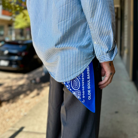 A person outdoors wearing a light blue and white vertically striped button-up shirt, dark gray trousers, and a bright blue bandana tucked into the back of the shirt. The bandana has white paisley patterns and the text 'OLDE SOUL BARBER'.