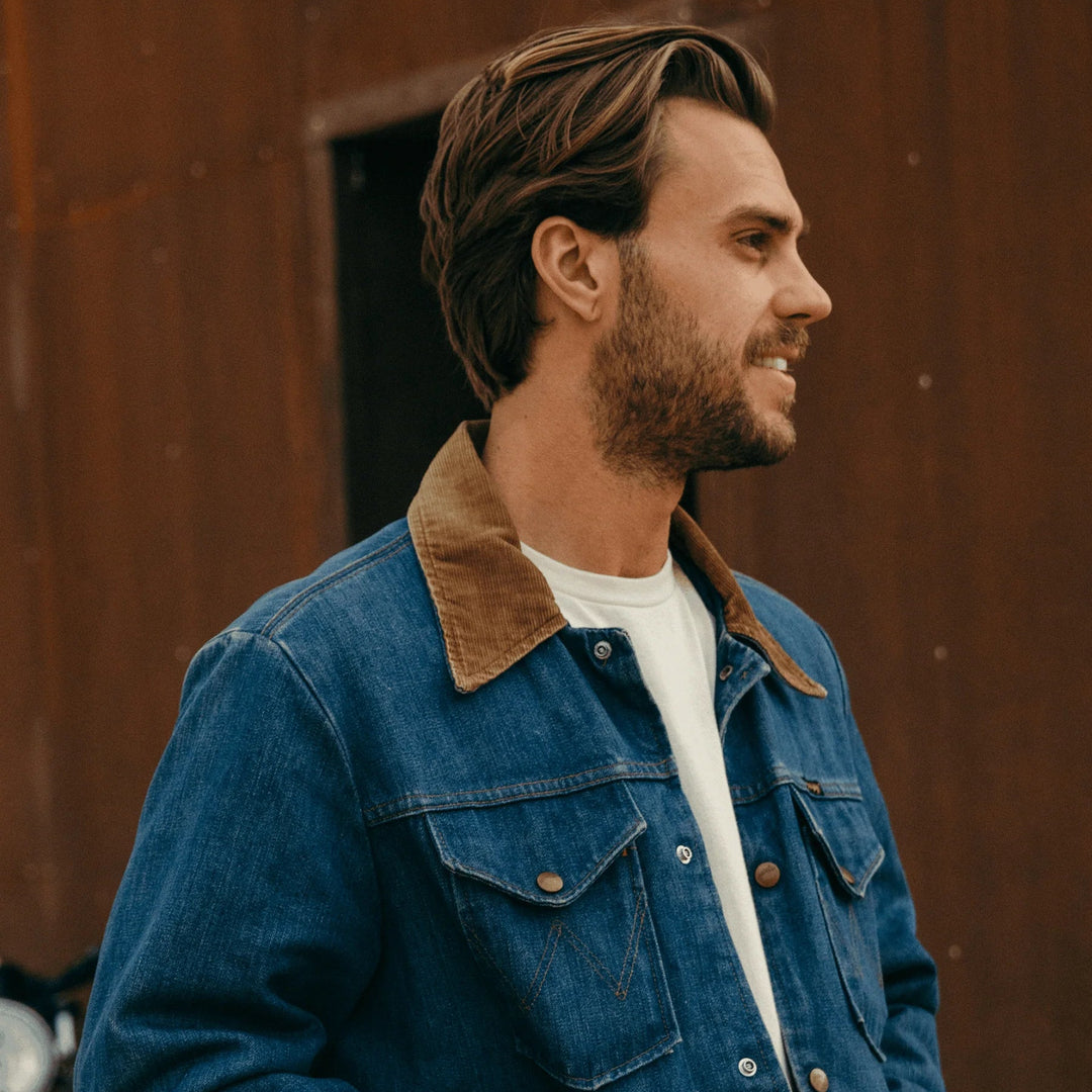 Man wearing a blue denim jacket with a brown collar against a rust-colored wall.
